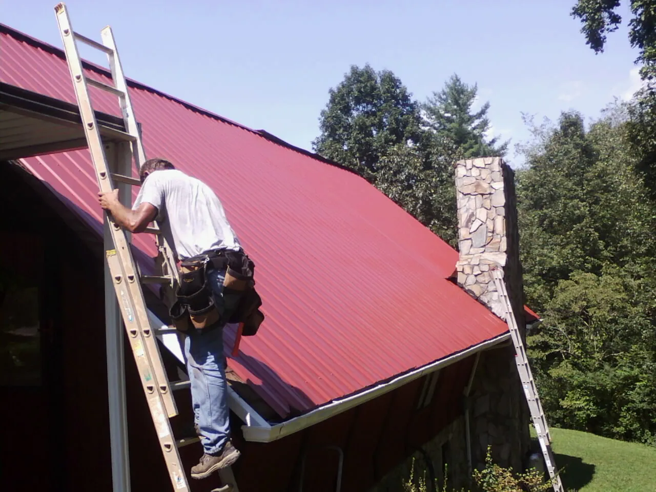 Roofer on a ladder beside a red metal roof and stone chimney — Soffit & Fascia Repair by Roofing Brothers in Northport